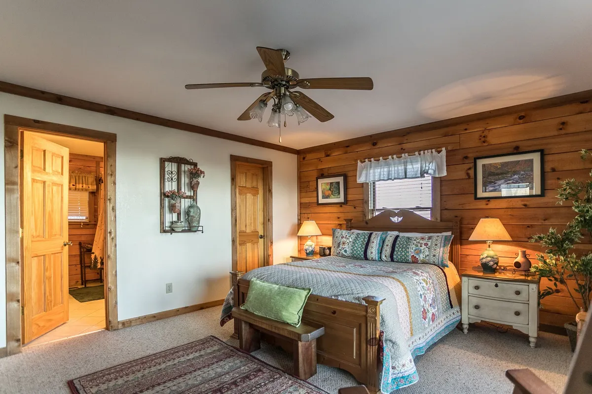 An open loft space with a colorful quilted bed, wooden walls, decorative lamps, and artwork, creating a comfortable and homey ambiance. The layout suggests a social sleep area ideal for groups or siblings, with an open view to the rest of the cabin below.