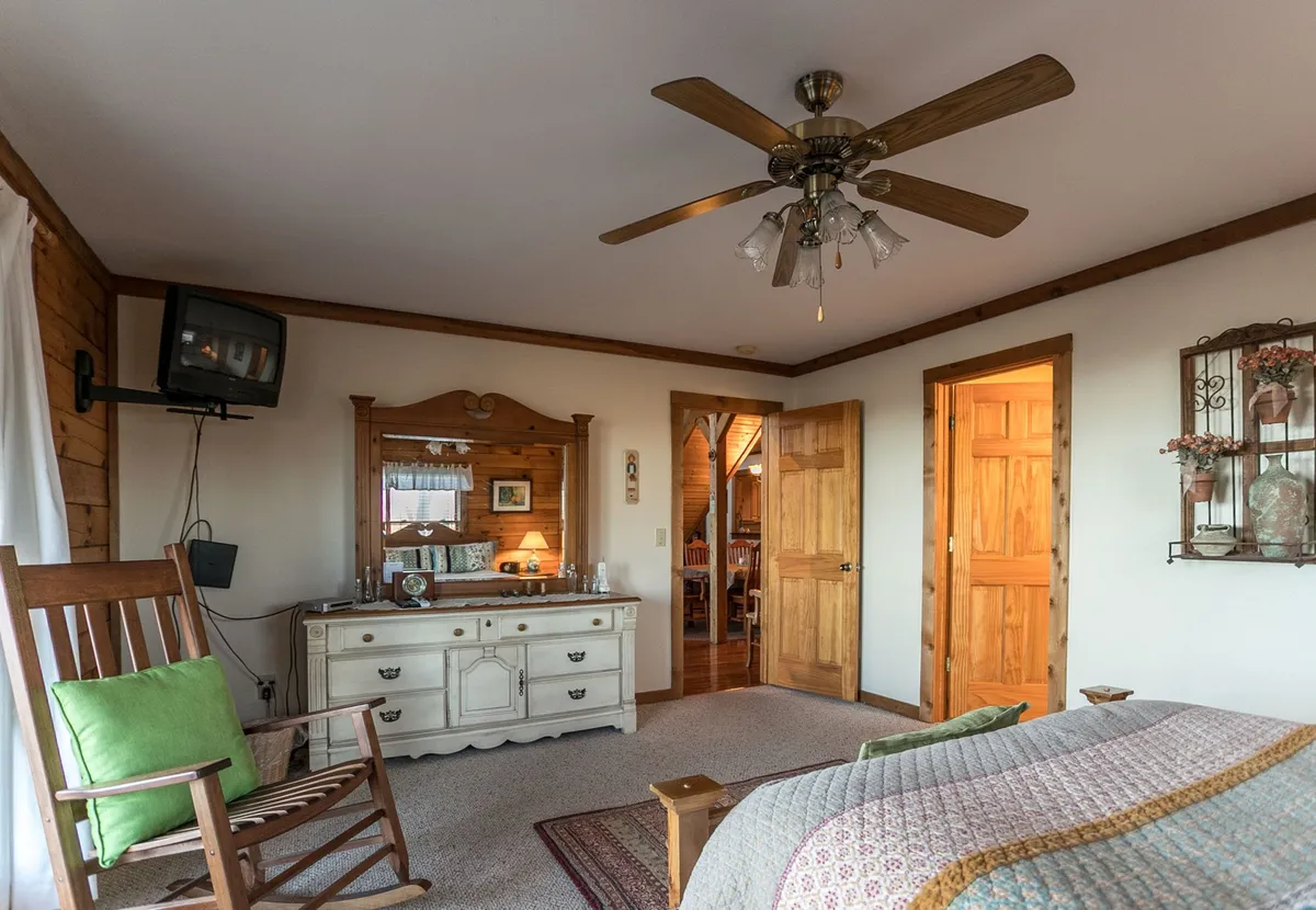 A sunlit bedroom featuring a comfortable queen bed, a dresser, and a cozy rocking chair positioned near wooden doors. The room is decorated with charming wall accents and has a TV, inviting relaxation. Soft natural light streams in, suggesting deck access and a tranquil morning atmosphere.