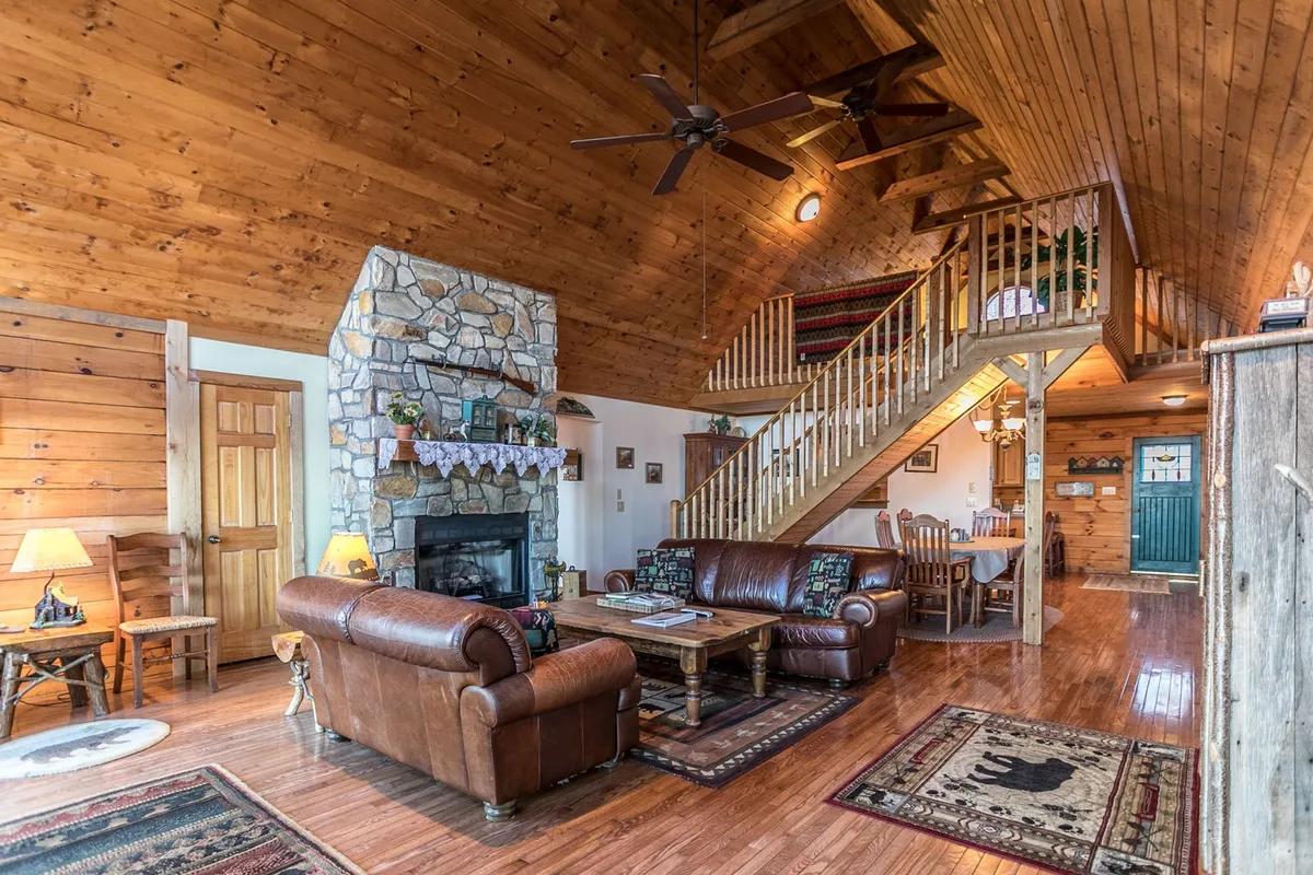 This image features a spacious, open-plan living area with a prominent stone-faced gas fireplace, leather sofas, a visible dining table, and a wooden staircase. Large windows allow natural light to pour in, creating a warm and inviting gathering space.