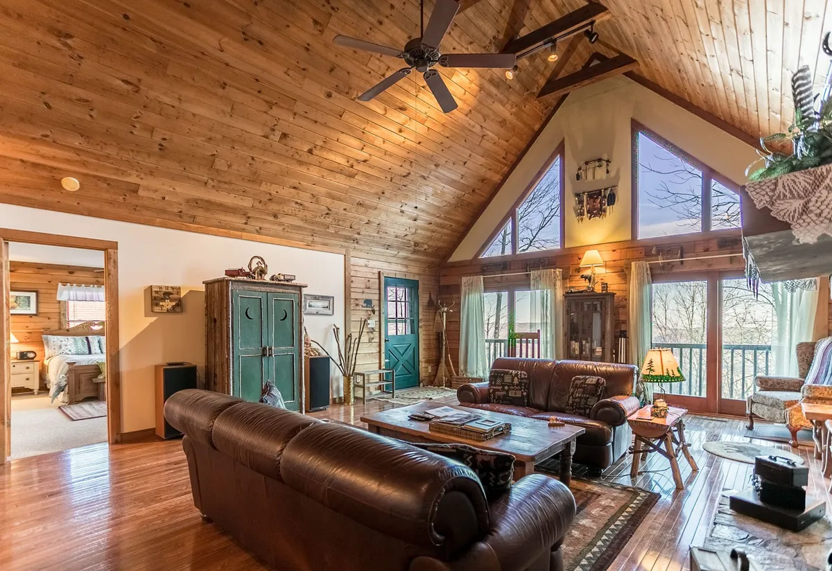 This image showcases a living room with leather sofas, a rustic coffee table, wooden beamed ceilings, and large windows framing scenic views. The space is bathed in natural light and exudes warmth and comfort.