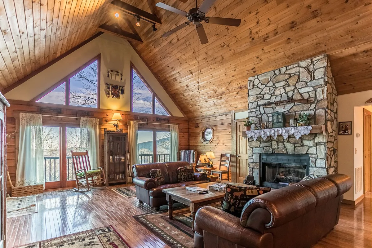 The image shows a cozy interior living space with a stunning floor-to-ceiling stone fireplace as the centerpiece. Large windows flood the room with natural light and frame breathtaking mountain views, while a warm wooden ceiling and plush leather sofas create an inviting spot for relaxing together. The scene suggests a welcoming environment perfect for family gatherings, enjoying a book by the fire, or watching the sunset through the windows.