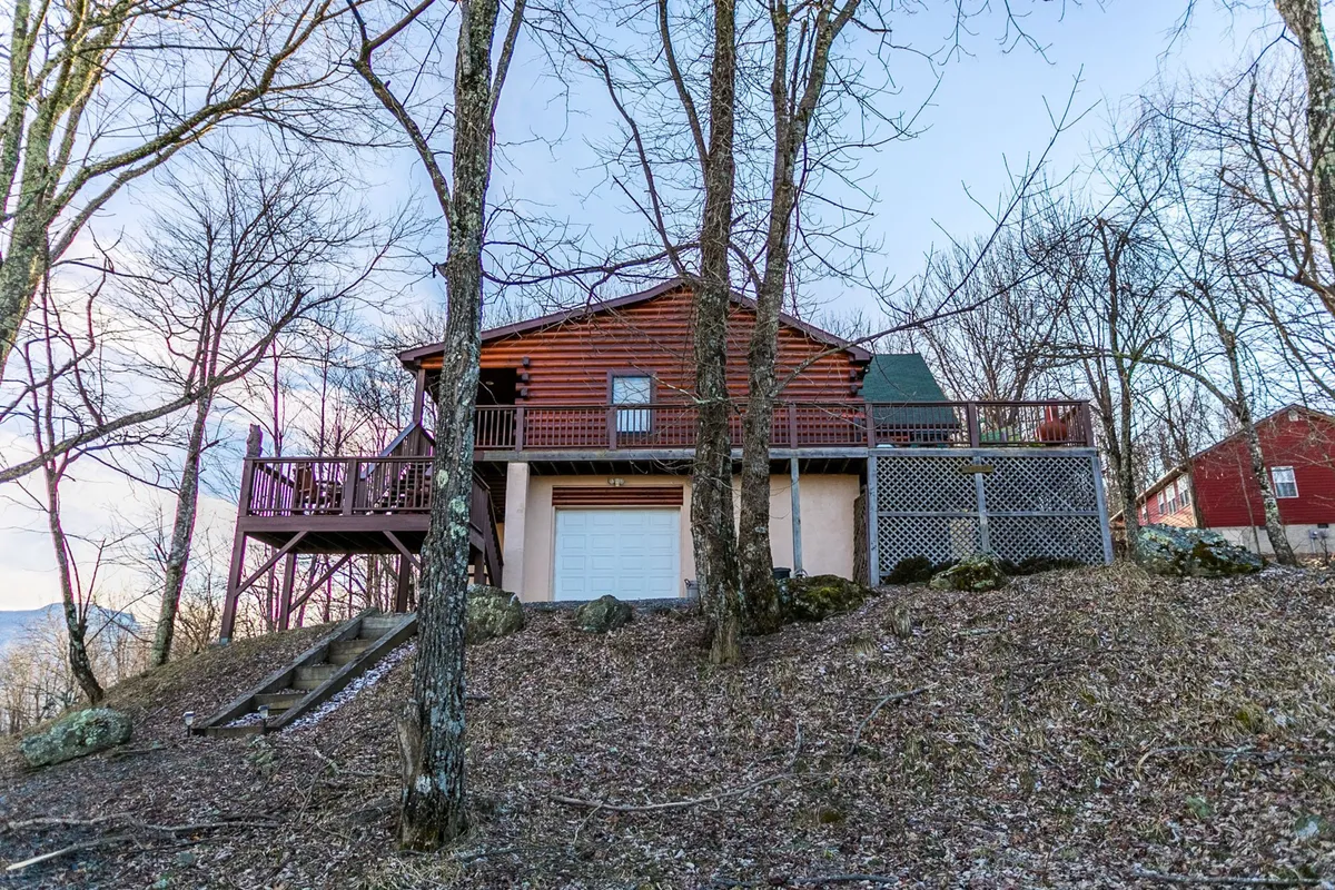 A wide view of the log cabin’s exterior, featuring the main deck and surrounded by mature trees. The inviting deck is shown as an ideal space for socializing, outdoor gatherings, or simply appreciating the peace of the mountain setting. The log construction reinforces both rustic charm and year-round livability.