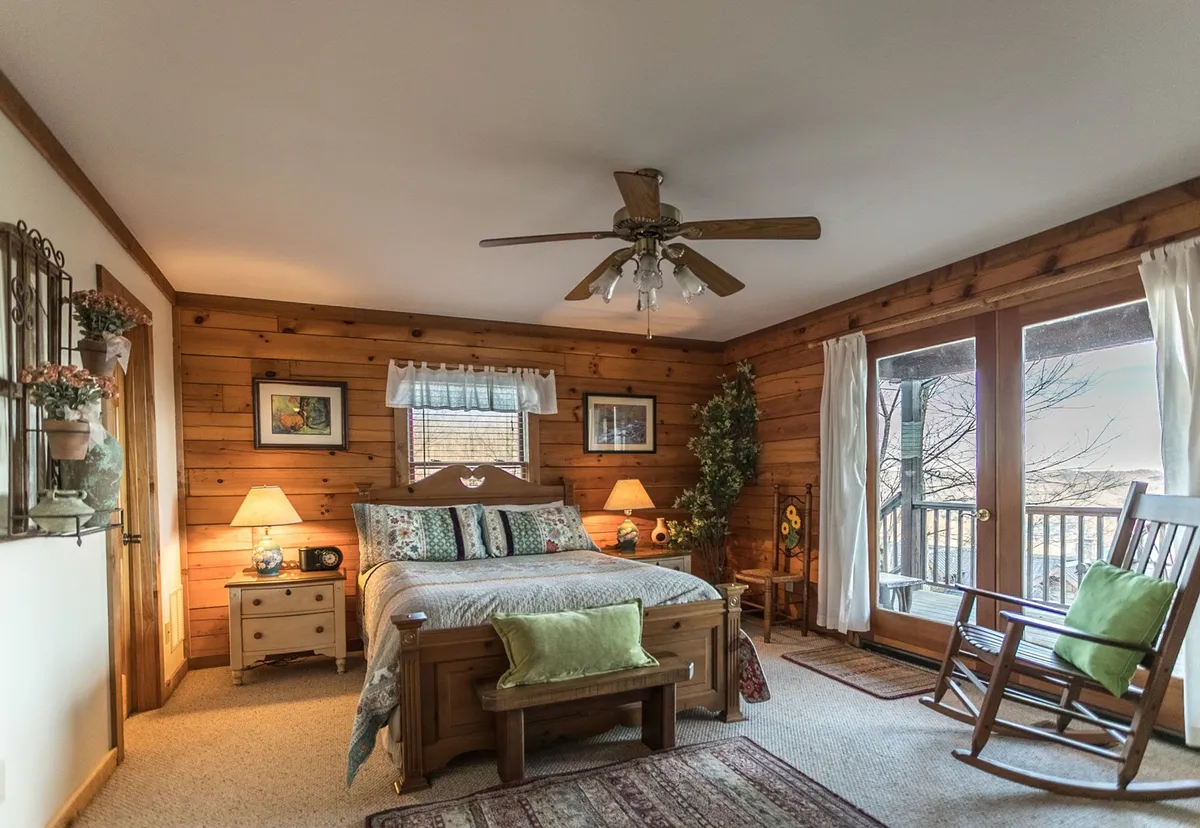 This inviting bedroom scene features a wooden bed, a cozy rocking chair next to large windows, table lamps, and touches of greenery. The expansive windows offer glimpses of the natural world outside, suggesting an easy transition from indoor comfort to outdoor exploration.