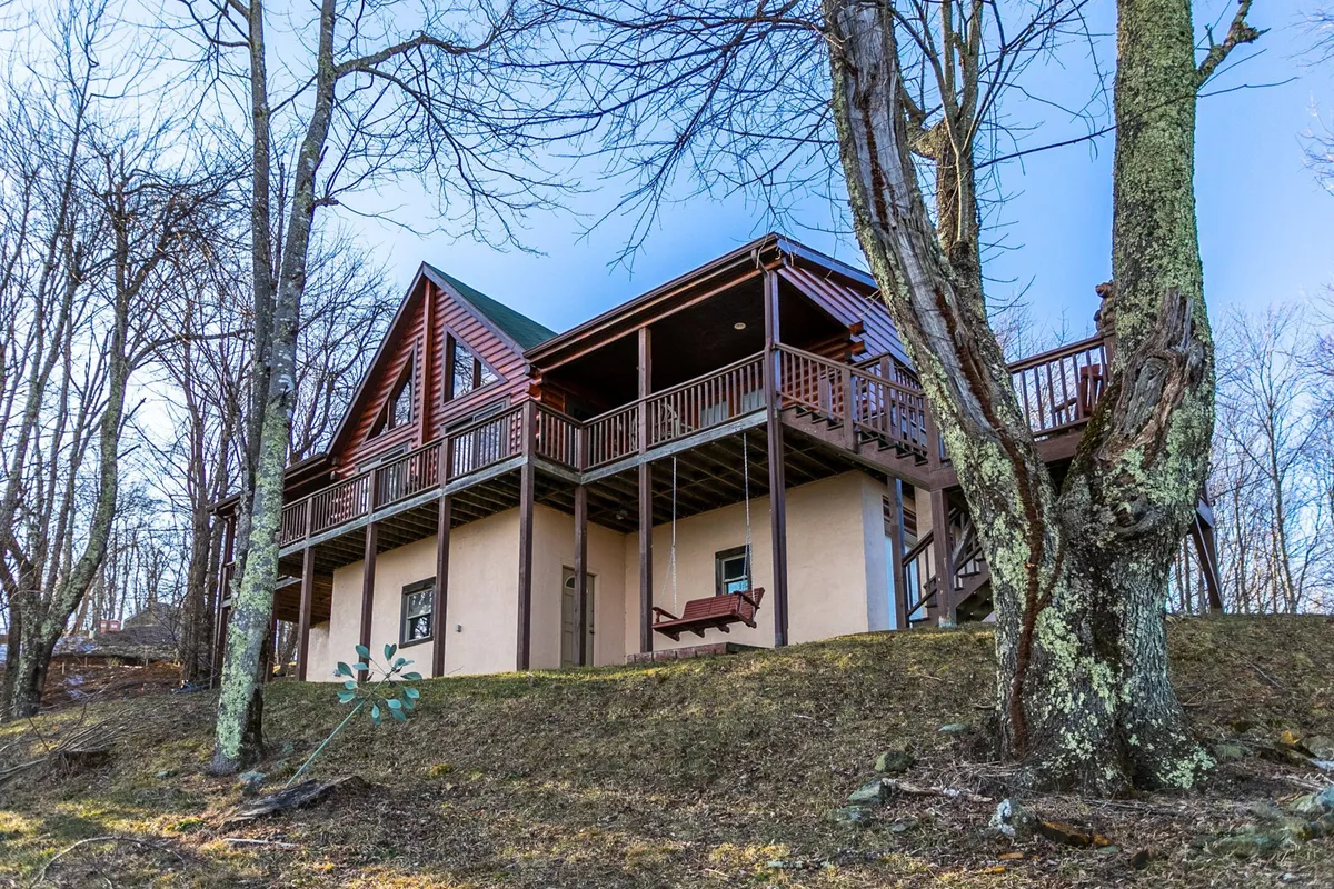 A spacious wooden balcony attached to the cabin, featuring a sloping roof, large windows, and nestled among lush trees. Family members are visible enjoying the space, some gathered for a barbecue while others relax and take in the mountain views. The scene captures the welcoming, communal feel of the deck, enhanced by the expansive natural backdrop.
