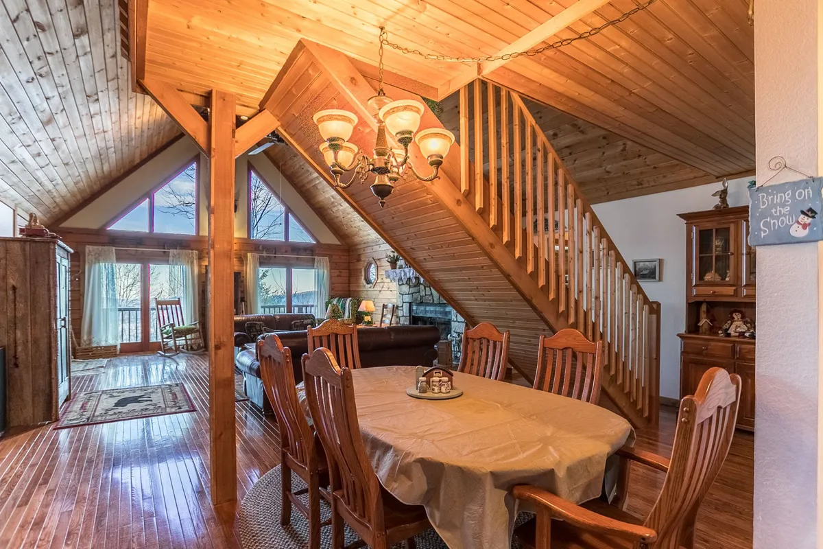 A spacious dining area with a large table set beneath a chandelier, adjacent to a staircase and bordered by expansive windows that fill the room with natural light. The setup is ideal for group meals, dinner parties, or game nights, offering plenty of space for everyone to gather comfortably.
